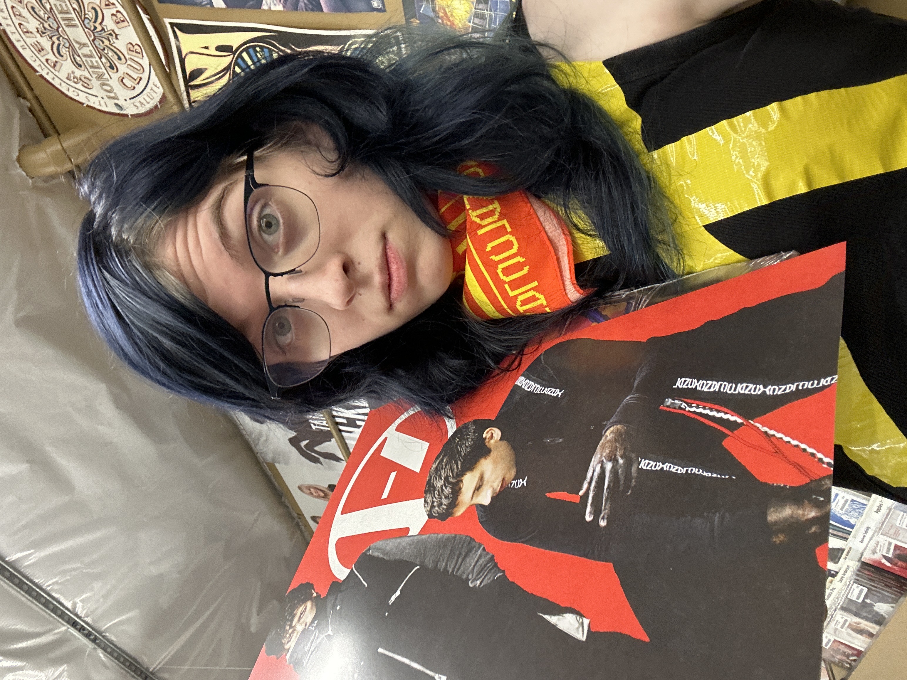 A photo of a girl with blackish blue hair holding a poster of Twenty One Pilots. She has yellow tape across her heart and a red and yellow bandana on. She's wearing glasses