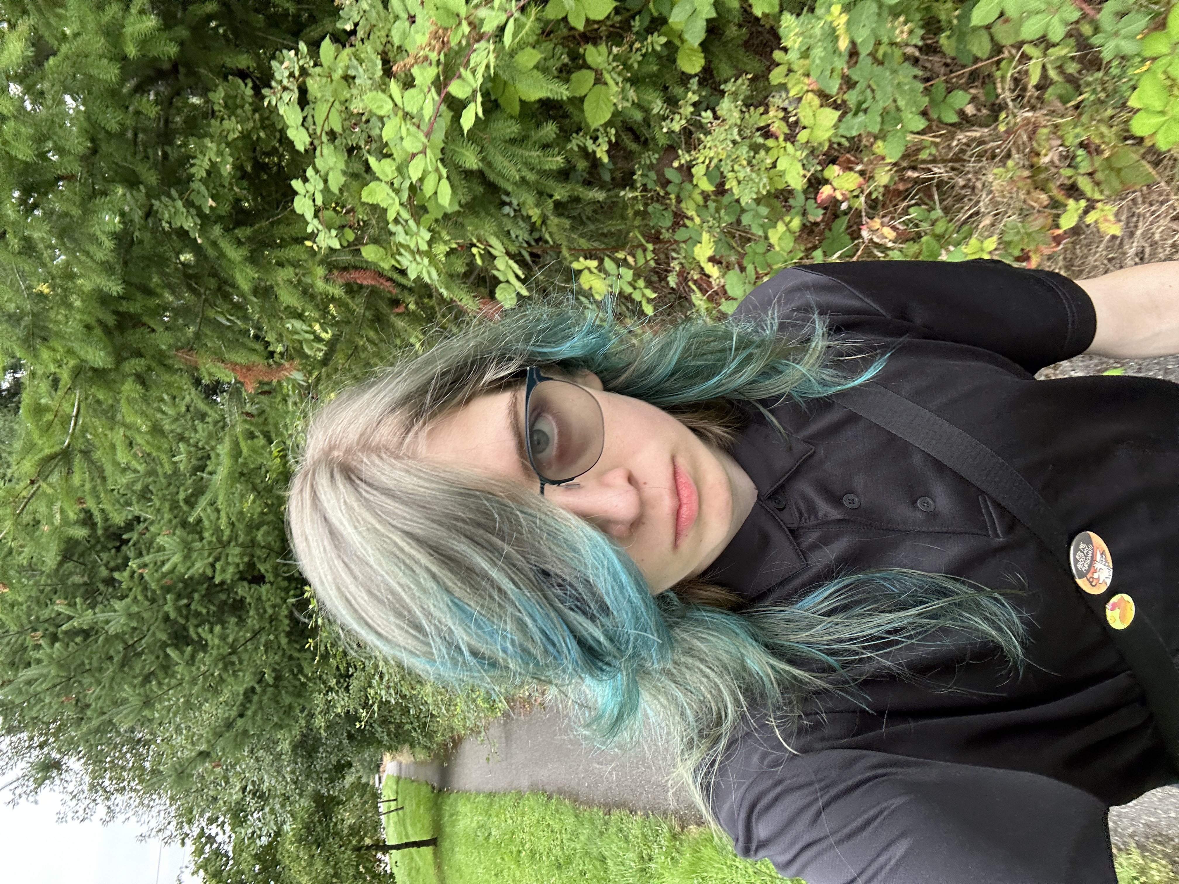 A selfie of a girl with blue hair staring intently into the camera. She's wearing a black dress shirt and has a bag slung across her chest.
